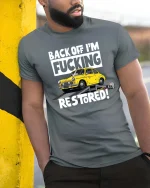 Back Off I’m Restoring Classic Car Mechanic Humor Tshirt - model wearing grey tee, leaning on column