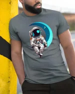 Astronaut Sitting On Moon Cute Space Graphic T-Shirt Design - model wearing grey tee, leaning on column